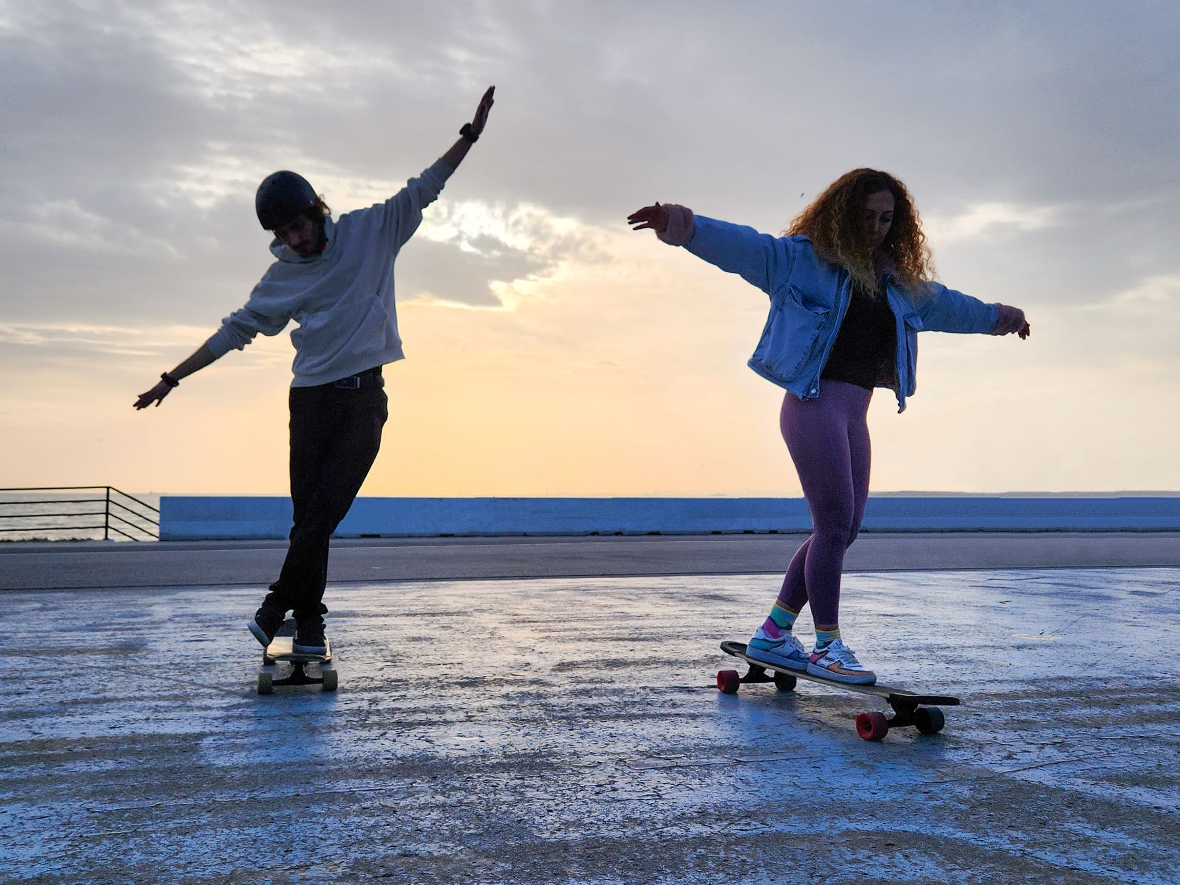 Two people skateboarding with their arms out on a seafront at sunset