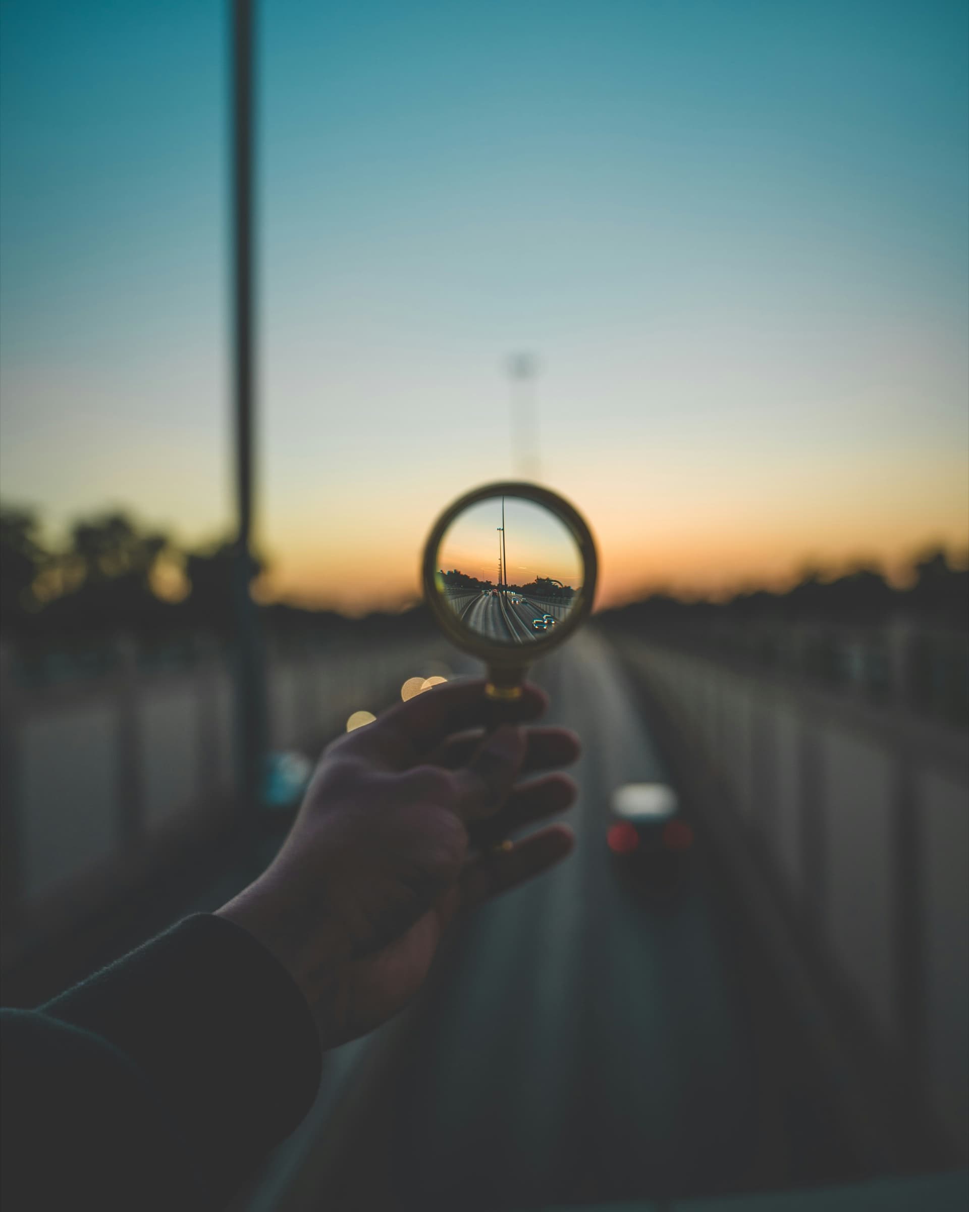 A hand holding a magnifying glass up to a road at sunset, bringing the far end into focus
