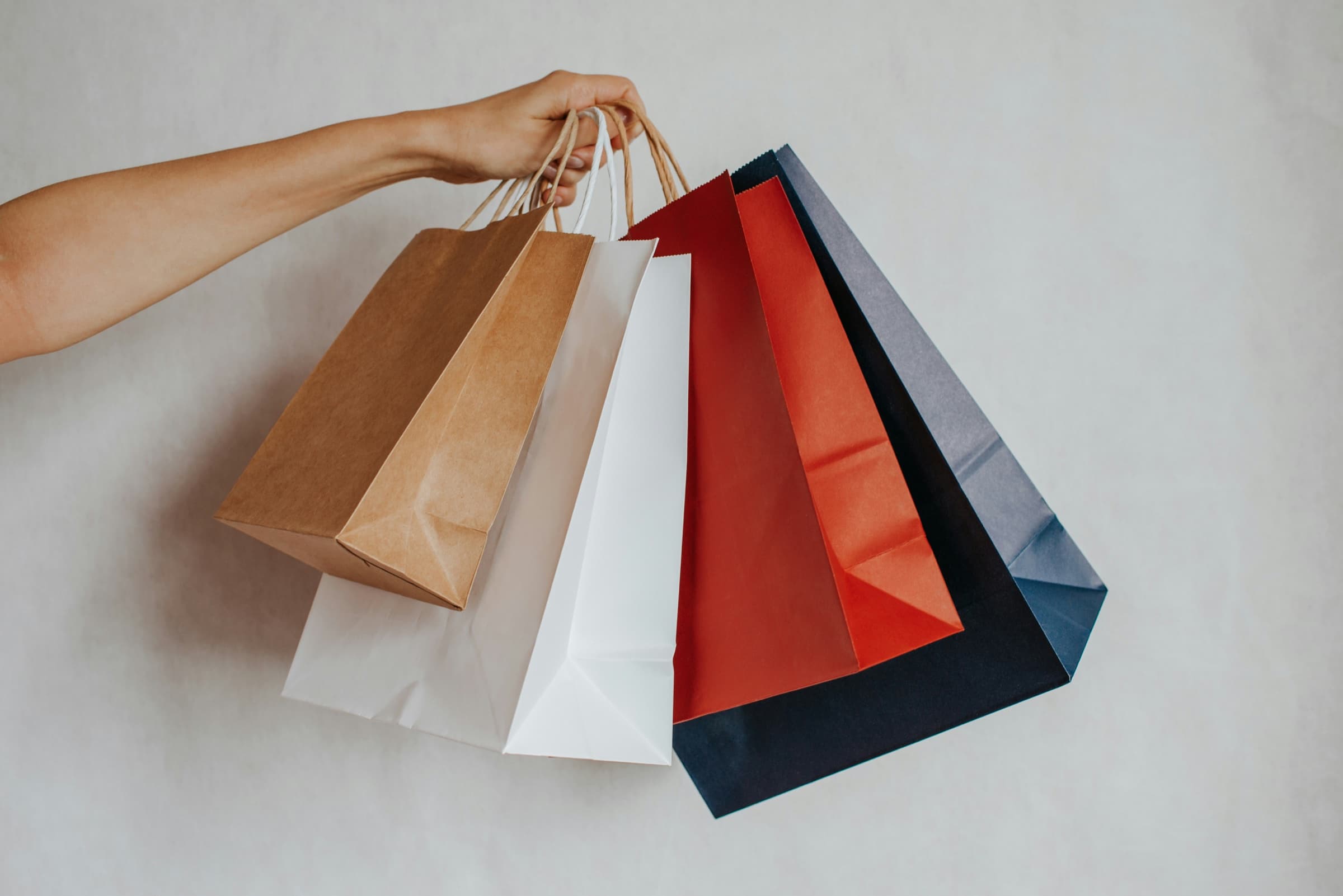 A hand holding up five paper shopping bags in different colours