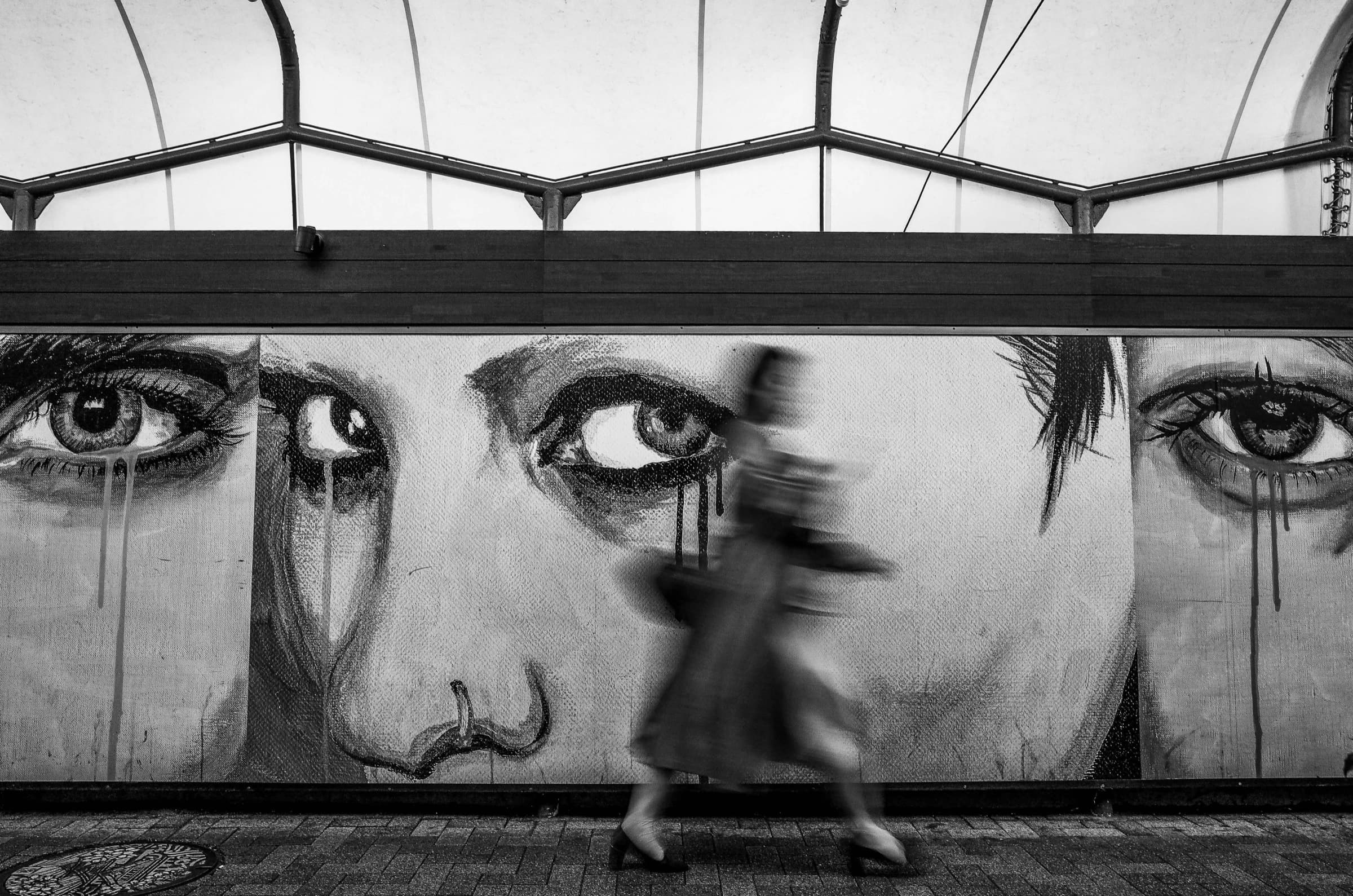A woman walking past a wall mural of large staring eyes, in black and white